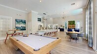 Located at 2800 Cheval Street in Orlando, FL, this expansive single-floor community clubhouse offers a generously proportioned open-plan interior designed for socializing and relaxation. The space is anchored by a grand living area with soaring ceilings and an impressive wall of arched and gridded windows that flood the room with natural light while framing lush outdoor views of the surrounding pool and greenery. Light wood-look flooring runs throughout, complementing the soft, neutral color palette of warm whites and grays.

The lounge area is elegantly furnished with cream curved accent chairs, navy blue wingback seating, and a central coffee table, all arranged atop layered area rugs. A striking living wall of lush greenery serves as a focal point near the reception area, paired with geometric navy cabinetry and a round decorative mirror. Tall French doors and patterned drapery panels add architectural character and connect the interior to the outdoors.

A well-appointed kitchen bar features a sleek white island with bar-height seating, stainless steel appliances, and pendant lighting over a wood-panel accent wall with a mounted display. Adjacent to the kitchen, a billiards table anchors a dedicated game area, while a separate dining nook with a round table provides additional gathering space. The overall ambiance is bright, modern, and inviting.