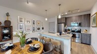 This contemporary single-floor residence offers a thoughtfully designed open-concept layout with modern finishes throughout. The kitchen is a standout, featuring dark wood-grain cabinetry, stainless steel appliances including a refrigerator, range, and over-the-range microwave, complemented by granite countertops and a mosaic tile backsplash. A large kitchen island with seating for multiple bar stools serves as both a functional workspace and a social hub, flowing seamlessly into the dining and living areas. Light wood-tone flooring runs throughout, adding warmth and continuity across the main living spaces. The entryway welcomes residents with clean lines and art-adorned walls, offering a glimpse into the kitchen just steps away. The living room is bright and inviting, with large sliding glass doors that open to a private balcony overlooking the surrounding development. The bedroom is a serene retreat, featuring dual windows that frame tranquil waterfront and treetop views, along with a ceiling fan for added comfort. Two full bathrooms are finished with large-format marble-style tile flooring, granite-topped dark wood vanities, soaking tubs with shower combinations, and wall-mounted storage cabinets, exuding a clean and polished aesthetic throughout.