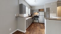 Located at 10 Kenley Way in Birmingham, Alabama, this single-floor home offers a cohesive and functional layout with modern finishes throughout. The kitchen is a standout feature, outfitted with gray shaker-style cabinetry, dark wood-look flooring, stainless steel appliances including an electric range and full-size refrigerator, and a double-basin sink beneath a bright window. A breakfast bar peninsula provides additional counter space and opens seamlessly into the main living area, which is bathed in natural light from large windows and a sliding glass door that leads to an outdoor balcony. The living space continues the warm wood-look flooring and benefits from a neutral gray color palette that lends a clean, contemporary feel. The bedrooms feature plush wall-to-wall carpeting, ceiling fans with light fixtures, generous windows with blinds, and ample closet access via a connecting hallway. The bathrooms are well-appointed — one features a wide vanity with extended countertop space, a large mirror, and a tub-shower combination with tiled surround, while another includes a sleek glass-door walk-in shower with white tile. Crown molding accents and textured ceilings add architectural character throughout this well-maintained home.