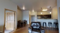 This single-floor space features an open-concept layout that seamlessly connects the living, dining, and kitchen areas into one cohesive environment. The living area is anchored by a dark leather armchair, positioned to take advantage of the open sightlines throughout the space. Warm hardwood-style flooring runs throughout, adding a sense of continuity and warmth to the neutral gray walls.

The dining area accommodates a rectangular wooden table with matching chairs, positioned near the living space and comfortably sized for everyday use. A doorway nearby provides access to an additional room or hallway, contributing to the functional flow of the layout.

The kitchen is situated along one wall and features dark cabinetry, granite-style countertops, and stainless steel appliances including a refrigerator and microwave. A raised breakfast bar with a metal barstool creates a casual seating option and serves as a natural visual divider between the kitchen and living areas. Pendant lighting hangs above the bar, complementing the flush-mount ceiling fixtures that provide consistent illumination throughout the space. The overall ambiance is practical, clean, and inviting.