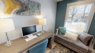 This thoughtfully appointed business center combines functionality with sophisticated interior design, creating a productive yet inviting workspace. The room is anchored by a wood-toned desk outfitted with a desktop computer, keyboard, and dual crystal-base table lamps that cast warm ambient light beneath a vibrant abstract painting in teal, gold, and gray tones. A teal upholstered rolling chair completes the workstation setup. Opposite the desk, a plush gray velvet chaise lounge is adorned with decorative pillows in teal, geometric gold, and neutral tones, positioned beside a window with wood blinds that allows natural light to filter in. A bold teal accent wall adds depth and color contrast to the otherwise soft gray walls, while light wood-tone flooring and an ornate area rug in navy, gold, and cream ground the space with warmth and texture. A striking decorative room divider composed of interlocking carved wood circles with mirrored panels serves as both a functional partition and an artistic focal point. Near the entry, a glass-panel interior door provides visual connectivity to adjacent spaces, and an HP multifunction printer atop a wood storage dresser ensures full office functionality. Crown molding and recessed lighting add refined architectural detail throughout.