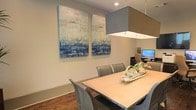 This single-floor space showcases a thoughtfully designed interior that blends modern aesthetics with comfortable functionality. The dining area serves as a central highlight, featuring a light wood rectangular table paired with studded gray upholstered chairs that add a touch of refined elegance. Overhead, a large square pendant light with a gray fabric shade provides focused, ambient illumination over the dining surface.

The walls are finished in a soft, neutral tone that complements the warm hardwood flooring below, creating a cohesive and inviting atmosphere throughout. A standout decorative feature includes a pair of large-scale abstract canvas artworks displayed prominently on the dining room wall. The paintings incorporate oceanic blues, whites, and sandy neutrals, lending the space an artistic, coastal-inspired character.

A natural centerpiece arrangement rests on the dining table, adding an organic element to the setting. In the background, a workstation area with a monitor is partially visible, suggesting the space may serve a dual purpose as both a dining and functional work environment. Recessed ceiling lighting further enhances the clean, contemporary feel of this well-appointed interior.