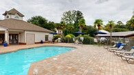 Located at 300 Palmetto Park Blvd in Lexington, South Carolina, this outdoor amenity area centers around a generously sized swimming pool with a classic freeform shape, offering both shallow and deeper sections accessible via stainless steel handrails. The surrounding deck is finished with attractive interlocking brick pavers, providing a durable and visually appealing surface throughout the space. Multiple seating areas are distributed around the pool, furnished with chairs and tables shaded by navy blue market umbrellas, ideal for relaxing poolside. A dedicated grilling station with a full-size gas grill and adjacent outdoor dining furniture offers a convenient space for outdoor cooking and entertaining. A pergola-style structure anchors one corner of the deck, adding architectural interest and additional shade. The pool area is enclosed by a classic wrought iron-style fence, ensuring safety while maintaining open sightlines to the surrounding greenery. Lush landscaping, including tall palm trees, ornamental shrubs, and decorative planters, lends a tropical ambiance to the setting. A single-story clubhouse or amenity building with a pitched roof, shuttered windows, and brick accents is situated directly adjacent to the pool, providing convenient access to facilities. The overall atmosphere is resort-like, offering a well-maintained and inviting outdoor retreat.