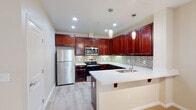 This single-floor residence offers a thoughtfully designed open-concept layout that seamlessly connects the living area to the kitchen. The living space features neutral-toned carpeting, smooth walls in warm beige tones, and natural light streaming in through windows with vertical blinds, creating a bright and welcoming atmosphere.

The kitchen is a standout feature, showcasing rich, deep-toned wood cabinetry that extends to the ceiling for generous storage. Light-colored quartz countertops provide a striking contrast against the dark cabinetry, while a mosaic tile backsplash in soft greens and grays adds texture and visual interest. Stainless steel appliances — including a refrigerator, electric range, and microwave — deliver a sleek, modern aesthetic. A peninsula with an undermount sink and bar seating bridges the kitchen and living area, ideal for casual dining or entertaining. Pendant lighting above the peninsula adds a refined finishing touch. Light wood-tone flooring grounds the kitchen space.

The bedroom is a serene retreat with plush carpeting, a window with natural light, and ample room for furnishings. The residence includes two full bathrooms: one featuring a soaking tub, a wide vanity mirror, and white cabinetry; and a second with a glass-enclosed walk-in shower, wood-tone flooring, and a matching white vanity — both finished in clean, neutral palettes.