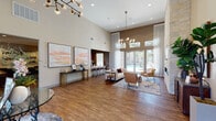 This single-floor communal lounge space is designed to impress, combining contemporary aesthetics with inviting functionality throughout its open layout. The main living area commands attention with soaring double-height ceilings and a striking floor-to-ceiling stacked stone fireplace wall, complete with a linear electric fireplace and a wall-mounted television flanked by symmetrical artwork. Warm hardwood-style flooring runs throughout, complementing the neutral wall tones and the rich textures of leather accent chairs, a plush sofa, and a layered area rug.

Sputnik-style globe chandeliers add a modern, artistic touch to the overhead space, while an abundance of natural light pours in through tall floor-to-ceiling windows and glass French doors that offer direct views of an outdoor pool area. Sheer white drapery softens the light and adds an element of elegance.

A dedicated lounge alcove near the entry features a console table and a pair of vibrant abstract paintings, providing a welcoming focal point. Adjacent to the main area, a fully equipped galley-style kitchenette offers rich mahogany cabinetry, quartz countertops, a stainless steel sink, a beverage refrigerator, and a wall-mounted TV — ideal for hosting and entertaining. A billiards table is also visible in a secondary activity area, further highlighting the space's versatility as a multi-functional gathering destination.