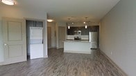 This single-floor space showcases a contemporary open-concept design that seamlessly integrates the living area and kitchen. The kitchen is a standout feature, equipped with stainless steel appliances including a gas range, microwave, and refrigerator. Dark espresso cabinetry contrasts elegantly with a light granite countertop island, which serves as both a prep space and a casual dining or gathering spot. A classic subway tile backsplash adds a clean, polished aesthetic, while pendant lighting and a track light fixture provide focused illumination throughout the kitchen area.

The flooring throughout the open living space consists of durable wood-look planks in a warm gray-brown tone, lending a cohesive and modern feel to the interior. A stacked washer and dryer unit is conveniently housed within the living area, offering in-unit laundry accessibility without requiring a dedicated utility room. The neutral wall tones and recessed lighting contribute to a bright, airy atmosphere. The overall design balances practicality with contemporary style, making the space both functional and visually appealing.