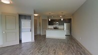 This single-floor space showcases a contemporary open-concept design that seamlessly integrates the living area and kitchen. The kitchen is a standout feature, equipped with stainless steel appliances including a gas range, microwave, and refrigerator. Dark espresso cabinetry contrasts elegantly with a light granite countertop island, which serves as both a prep space and a casual dining or gathering spot. A classic subway tile backsplash adds a clean, polished aesthetic, while pendant lighting and a track light fixture provide focused illumination throughout the kitchen area.

The flooring throughout the open living space consists of durable wood-look planks in a warm gray-brown tone, lending a cohesive and modern feel to the interior. A stacked washer and dryer unit is conveniently housed within the living area, offering in-unit laundry accessibility without requiring a dedicated utility room. The neutral wall tones and recessed lighting contribute to a bright, airy atmosphere. The overall design balances practicality with contemporary style, making the space both functional and visually appealing.