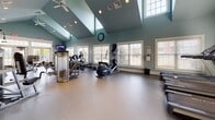 Located at 1085 Hartford Tpke in Vernon Rockville, Connecticut, this fitness center offers a spacious, single-floor layout designed to support a full range of workout activities. The facility is immediately distinguished by its dramatic vaulted ceilings, which lend an open, airy quality to the entire space. Painted in a calming blue-gray palette, the walls complement the light-toned flooring and white trim throughout, creating a clean and welcoming environment.

Natural light pours in through multiple large windows and elevated clerestory windows, brightening the interior throughout the day. French doors along one wall provide both additional light and convenient access. Recessed ceiling lights and ceiling fans supplement the natural light and ensure comfortable airflow during workouts.

The main exercise area is generously proportioned and well-organized, housing a diverse selection of equipment. Cardio machines include ellipticals, stationary bikes, and additional aerobic equipment, while strength training is supported by Nautilus cable machines and various weight benches. Stability balls and free weights round out the offerings, catering to a broad spectrum of fitness routines.

A wall-mounted television keeps members entertained, and a coat hook area near the entry provides practical convenience. The space also connects to an adjacent hallway, offering easy access to additional amenities within the building.