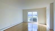 This single-floor space offers a clean and functional layout with a bright, open feel. The main living area is finished with polished hardwood floors that reflect natural light beautifully, enhancing the sense of space and warmth throughout. Neutral, white-toned walls keep the interior feeling fresh and versatile.

A standout feature of the space is the sliding glass door, which opens directly onto a private balcony with a metal railing. The door allows abundant natural light to fill the room while providing pleasant views of a grassy outdoor area and surrounding greenery. The balcony offers a comfortable outdoor extension of the living space.

Along the base of one wall, a baseboard heating unit provides efficient climate control, a practical and unobtrusive feature well-suited for colder climates. The overall design is understated and move-in ready, with a straightforward layout that accommodates a range of furniture arrangements. The combination of natural light, outdoor access, and warm hardwood flooring gives the space an inviting and comfortable ambiance.