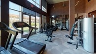 This fitness center is designed to provide a comprehensive workout environment within a sleek, modern setting. The space is organized to accommodate a wide range of exercise needs, beginning with a welcoming entry area finished with warm wood-toned flooring, branded area rugs, and wall-mounted media screens. A neatly arranged free weight section near the entrance includes a full dumbbell rack and a medicine ball tower, catering to functional and strength training routines.

The main workout floor transitions to a patterned carpet surface and features an extensive lineup of cardio equipment, including treadmills, stationary bikes, and elliptical machines positioned along a wall of large, floor-to-ceiling windows. These expansive glazed panels flood the space with natural light and provide open views of the surrounding landscape, creating an energizing atmosphere during workouts.

Structural columns punctuate the interior, while dark wood wall paneling and large wall mirrors enhance the sense of depth and add a polished aesthetic. The ceiling rises notably, contributing to an open, airy feel. Strength training machines and cable systems are arranged throughout the space, ensuring a well-rounded equipment offering. Recessed and track lighting complement the natural light, maintaining consistent illumination throughout.