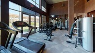 This fitness center is designed to provide a comprehensive workout environment within a sleek, modern setting. The space is organized to accommodate a wide range of exercise needs, beginning with a welcoming entry area finished with warm wood-toned flooring, branded area rugs, and wall-mounted media screens. A neatly arranged free weight section near the entrance includes a full dumbbell rack and a medicine ball tower, catering to functional and strength training routines.

The main workout floor transitions to a patterned carpet surface and features an extensive lineup of cardio equipment, including treadmills, stationary bikes, and elliptical machines positioned along a wall of large, floor-to-ceiling windows. These expansive glazed panels flood the space with natural light and provide open views of the surrounding landscape, creating an energizing atmosphere during workouts.

Structural columns punctuate the interior, while dark wood wall paneling and large wall mirrors enhance the sense of depth and add a polished aesthetic. The ceiling rises notably, contributing to an open, airy feel. Strength training machines and cable systems are arranged throughout the space, ensuring a well-rounded equipment offering. Recessed and track lighting complement the natural light, maintaining consistent illumination throughout.