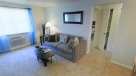 This single-floor space features a living room designed for comfort and functionality. The room is finished with wall-to-wall carpeting in a warm neutral tone, contributing to a relaxed and inviting atmosphere. The walls are painted in a soft, light gray-blue hue, creating a calm backdrop throughout the space.

The living area is centered around a plush gray sofa adorned with a mix of decorative pillows in dark, patterned, and muted yellow tones. A rectangular black-framed mirror is mounted on the wall above, adding depth and visual interest. A floor lamp with a classic white shade provides warm ambient lighting, while a small side table sits nearby, holding decorative branches that add a natural, organic touch.

A low, oval-shaped black coffee table anchors the seating area and features candle holders and reading material on its surface. Blue sheer curtains frame a window on the left wall, allowing soft natural light to filter into the room.

An open doorway on the right leads to an adjoining room, suggesting a well-connected layout that promotes easy flow between spaces. The overall ambiance is tidy, understated, and functional.