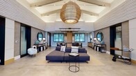 Located at 15200 Leicestershire St in Woodbridge, Virginia, this single-floor amenity center is thoughtfully designed to blend modern aesthetics with functional comfort. The grand lobby immediately impresses with soaring vaulted ceilings accented by exposed white beams and a striking oversized woven pendant light. Textured stone-clad walls and large grid-paned windows frame a spacious seating area anchored by a generous sectional sofa, creating an inviting communal atmosphere. Multiple lounge areas throughout the space continue this refined aesthetic, featuring layered pendant lighting, natural stone accent walls, and curated furnishings that balance warmth with contemporary style. The fully equipped fitness center is divided into distinct zones, including a cardio area lined with treadmills and cycling machines backed by floor-to-ceiling mirrors, and a dedicated strength training room with large windows that allow abundant natural light. A sleek, modern kitchen features dark cabinetry, white quartz countertops, patterned terrazzo-style tile flooring, stainless steel appliances, and an overhead grid of track lighting. A well-appointed pet spa offers a utility sink, open shelving, wood-paneled walls, and decorative tile floors. Dual drinking fountains with dark tile accents and glass-framed artwork further illustrate the attention to detail found throughout this exceptional amenity space.
