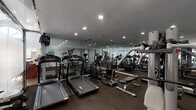 This fitness center is organized across a single floor and divided into two distinct workout zones separated by a floor-to-ceiling frosted glass partition with a chrome frame. The main exercise area features dark, textured carpet flooring and is lined with full-length mirrors along one wall, visually expanding the space and allowing users to monitor their form. Recessed lighting throughout provides consistent, even illumination across the entire facility.

The cardio zone is well-stocked with multiple treadmills, including NordicTrack TM6000 models, elliptical trainers, and stationary bikes, several equipped with individual monitors. Strength training equipment includes a multi-station cable machine, a weight bench, and a rack of dumbbells arranged in ascending order. A rowing machine and additional resistance equipment provide further workout variety.

The secondary zone, visible through the glass partition, features hardwood-style flooring and additional equipment, with a vaulted ceiling and fan overhead offering an open, airy atmosphere. Fitness center policies are posted prominently near the glass entry. The overall design is clean and functional, with a neutral color palette of grays and light blues contributing to a focused, professional training environment.