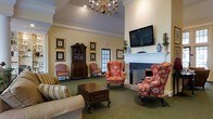 This single-floor space exudes a traditional, residential charm through its carefully curated interior design and inviting layout. The main gathering area serves as the heart of the space, centered around a classic brick fireplace with a crisp white mantel, above which a wall-mounted flat-screen television is thoughtfully installed. A chandelier with candelabra-style lighting hangs from the elevated ceiling, casting warm light throughout the room and complementing the elegant crown molding that frames the upper walls.

Seating is arranged to encourage conversation and comfort, featuring floral-upholstered wingback chairs and a plush striped sofa surrounding a dark wood coffee chest that doubles as a decorative accent table. The rich green carpet grounds the room with a sense of warmth and tradition. Along the walls, a stately dark wood secretary cabinet with glass-front display shelving adds a distinguished, antique character, while a collection of gold-framed botanical and decorative artwork adorns the walls.

Natural light filters in through a paneled glass door, providing a connection to the outdoors. Decorative accessories, including blue-and-white ceramic vases and potted greenery, complete the polished aesthetic. The overall ambiance is refined, cozy, and thoughtfully designed to feel both functional and welcoming.