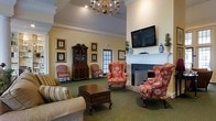 This single-floor space exudes a traditional, residential charm through its carefully curated interior design and inviting layout. The main gathering area serves as the heart of the space, centered around a classic brick fireplace with a crisp white mantel, above which a wall-mounted flat-screen television is thoughtfully installed. A chandelier with candelabra-style lighting hangs from the elevated ceiling, casting warm light throughout the room and complementing the elegant crown molding that frames the upper walls.

Seating is arranged to encourage conversation and comfort, featuring floral-upholstered wingback chairs and a plush striped sofa surrounding a dark wood coffee chest that doubles as a decorative accent table. The rich green carpet grounds the room with a sense of warmth and tradition. Along the walls, a stately dark wood secretary cabinet with glass-front display shelving adds a distinguished, antique character, while a collection of gold-framed botanical and decorative artwork adorns the walls.

Natural light filters in through a paneled glass door, providing a connection to the outdoors. Decorative accessories, including blue-and-white ceramic vases and potted greenery, complete the polished aesthetic. The overall ambiance is refined, cozy, and thoughtfully designed to feel both functional and welcoming.