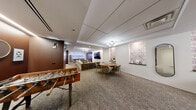 Located at 200 E Chestnut St in Chicago's vibrant Streeterville neighborhood, this well-appointed amenity lounge spans a generous single floor and is thoughtfully designed for relaxation and socializing. The space is divided into several distinct functional zones that flow seamlessly into one another.

The main living area serves as the centerpiece, featuring a large sectional sofa in warm taupe upholstery arranged around a wall-mounted flat-screen television, complemented by a pair of sleek round coffee tables and a striking multi-globe brass chandelier overhead. Recessed tray ceiling detailing adds architectural character to the room. Adjacent to this is a secondary lounge area with deep-toned rich brown accent walls, a curated gallery of framed artwork, and pairs of plush olive green armchairs — creating an intimate conversation nook.

A versatile gathering zone includes a round wood dining table with green velvet chairs and a decorative pendant light, alongside a seating vignette with burgundy accent chairs set against botanical-patterned wall panels. A foosball table occupies a dedicated game area, adding a playful dimension to the amenity space.

A bar-height table with gold-accented stools anchors a transitional corridor, while a staircase with metal railings connects to adjacent levels. The space is finished with a combination of neutral tile and patterned carpet flooring throughout, creating a polished and cohesive aesthetic.
