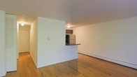 This single-floor space features a clean, open layout centered around warm hardwood flooring that runs throughout the main living area and hallway. The living space is generously proportioned, offering a blank canvas with neutral white walls and a flush-mount ceiling light that provides even illumination across the room.

A partial wall divides the kitchen from the living area, incorporating a pass-through counter with a dark countertop surface. The kitchen is equipped with dark wood cabinetry and a stainless steel refrigerator visible through the opening, creating a functional yet connected layout that allows interaction between the two spaces without fully opening them. A hallway extends from the main living area, suggesting additional rooms beyond the visible space.

Climate control is provided by a baseboard heating unit along the living room wall, a practical and common feature in residential units of this style. The overall aesthetic is straightforward and utilitarian, with an emphasis on usable space and easy maintenance. The neutral palette and hardwood floors provide a versatile foundation well-suited for a range of furnishing styles.