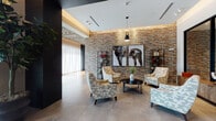 Located at SW 183rd Street in Palmetto Bay, Florida, this single-floor communal space offers a thoughtfully designed environment that balances modern architecture with warm, eclectic interiors. The main lobby welcomes visitors with large-format geometric-patterned tile flooring that transitions into rich hardwood, complemented by floor-to-ceiling sheer drapery that filters abundant natural light and frames views of lush Florida greenery outside. Structural columns clad in concrete-finish panels add an industrial-contemporary edge, while linear LED lighting overhead creates a clean, polished ambiance. A dedicated mail and service alcove sits conveniently off the entry corridor, appointed with dark cabinetry and framed artwork. The lounge area is a standout, featuring a dramatic exposed brick accent wall adorned with bold animal-themed artwork, surrounded by patterned accent chairs and a sleek round coffee table. A secondary lounge space showcases sculptural pendant lighting with woven, sun-burst designs, a deep sectional sofa, geometric-patterned rugs, and curated wall art — all opening toward an outdoor mural visible through full-height glass doors. Together, these spaces reflect a cohesive, design-forward aesthetic that is both inviting and visually dynamic.