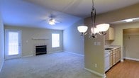 This single-floor space presents an open-concept layout that seamlessly connects the living and dining areas, creating a cohesive and functional environment. The living area is anchored by a tiled fireplace with a classic white mantel, serving as a natural focal point and adding warmth to the room. A ceiling fan overhead provides practical air circulation, while neutral-toned carpet throughout lends a soft, comfortable feel underfoot. Natural light filters in through a window positioned near the fireplace, brightening the space with a clean, airy quality. The walls are finished in a soft, warm beige tone that complements the light carpet and white trim detailing. Transitioning toward the dining area, an elegant multi-arm chandelier with frosted glass shades hangs overhead, providing both decorative appeal and ample illumination. A half-wall partition subtly defines the dining space from the kitchen area, where white cabinetry and an appliance are partially visible, hinting at a practical and well-appointed kitchen. The overall ambiance is calm, neutral, and move-in ready, with clean lines and understated finishes that provide a versatile backdrop for a variety of interior design styles.