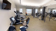 This fitness center offers a thoughtfully organized, single-floor exercise facility designed to support a full range of workout routines. The space is bright and open, with multiple windows along the walls allowing ample natural light to fill the room, complemented by overhead fluorescent lighting for consistent illumination throughout.

The cardio section is generously stocked with a lineup of elliptical trainers, treadmills, and a stationary bike, all positioned to take advantage of the natural light from the windows. The machines feature integrated digital displays, supporting a variety of workout programs and performance tracking.

On the opposite side of the floor, a dedicated strength training area provides cable and resistance machines suited for a range of muscle groups, offering users a complete gym experience without leaving the facility. Additional equipment, including a stability ball and exercise mat, is available for stretching and functional training.

The flooring throughout is a durable, low-pile carpet appropriate for a fitness environment, and the neutral-toned walls contribute to a clean, focused atmosphere. The layout is practical and spacious, allowing users to move freely between equipment. Overall, this fitness center is well-appointed and functional, catering to a variety of fitness needs and experience levels.