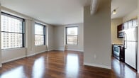 This single-floor urban residence combines classic architectural character with modern finishes throughout its thoughtfully laid-out rooms. The main living area is a standout, featuring richly stained hardwood floors that reflect natural light pouring in through multiple large, black-framed windows. These windows frame impressive views of the surrounding cityscape, including what appears to be a prominent domed landmark building — a rare and eye-catching feature. The neutral, warm-toned walls complement the warm flooring and create a bright, inviting atmosphere. Exposed ductwork along the upper walls adds an industrial loft-like character to the space. The bedroom offers plush carpeting underfoot and its own large window overlooking a vibrant urban street scene, providing both comfort and connectivity to the city below. The kitchen is equipped with stainless steel appliances, including a Frigidaire electric range, over-range microwave, and refrigerator, paired with dark cabinetry and clean white countertops. The bathroom features light tile flooring, a full bathtub and shower combination with white tile surround, a dark-toned vanity with a white countertop, and clean, functional fixtures throughout. Overall, this space offers a polished and comfortable urban living environment.
