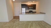 This single-floor residence offers a thoughtfully designed open-concept layout that seamlessly connects the kitchen and living areas, creating a sense of spaciousness and ease of movement throughout. The kitchen is a standout feature, outfitted with rich dark espresso cabinetry that provides ample storage and a striking visual contrast against the light granite countertops. A peninsula with a matching granite surface extends the workspace and doubles as a casual dining or entertaining area. Stainless steel appliances — including a refrigerator, range, and built-in microwave — lend a contemporary, polished finish to the space. A stainless steel sink is neatly integrated into the peninsula counter. Track lighting overhead ensures the kitchen is well-lit and functional. The adjoining living area is finished with neutral wall-to-wall carpeting that adds warmth and comfort underfoot, complementing the overall neutral color palette of the space. The layout flows naturally toward a private entry door, offering a welcoming transition point. The clean lines, modern finishes, and cohesive design throughout make this a practical and visually appealing single-level residence.