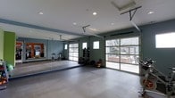 This single-floor fitness and yoga studio is thoughtfully designed to support a wide range of workout activities. The space is anchored by a polished concrete floor that provides a durable, easy-to-maintain surface suited for both high-intensity training and mindful movement practices. Large glass-panel garage doors span one wall, flooding the interior with natural light and offering open views of the surrounding outdoor area, creating an airy and energizing atmosphere.

A full-length mirrored wall on one side expands the sense of space while allowing users to monitor their form during exercise. The walls are painted in a deep teal-green tone, lending the studio a focused and modern aesthetic. Recessed ceiling lighting supplements the abundant natural light, ensuring the space remains well-lit at all times.

Equipment is neatly organized throughout the studio, including a power rack suitable for strength training, a dedicated dumbbell and weight plate storage rack stocked with a range of loads, and multiple cycling bikes for cardio workouts. A yoga studio area is visible through the mirrored wall, signaling a dedicated zone for stretching and mindfulness. Additional windows along the side walls further enhance ventilation and light. This studio offers a comprehensive, professionally appointed environment for fitness and wellness.