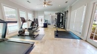 The Gatsby Fitness Center in Montgomery, Alabama is a thoughtfully designed, single-floor exercise facility that combines functionality with an elegant aesthetic. The space features polished travertine tile flooring throughout, accented by protective rubber mats positioned beneath the equipment. Crown molding and neutral-toned walls lend a refined, upscale character to the environment.

Natural light is a defining feature of this fitness center, with large windows dressed in white plantation-style blinds lining multiple walls. French doors provide both accessibility and an additional source of daylight, offering views of the surrounding outdoor area. A ceiling fan with integrated lighting ensures comfortable air circulation during workouts.

The facility is comprehensively equipped to support a full range of training needs. Cardio options include a treadmill, recumbent bike, upright bike, elliptical trainer, and a spin bike. A multi-station cable machine with an adjustable bench supports strength and resistance training, while a fully stocked dumbbell rack provides a wide selection of free weights for varied exercises. A large wall-mounted mirror visually expands the space and allows for form monitoring during workouts. A media console with a display screen rounds out the amenities, enhancing the overall workout experience. The Gatsby Fitness Center offers a polished and well-appointed environment suited for a comprehensive fitness routine.