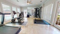 The Gatsby Fitness Center in Montgomery, Alabama is a thoughtfully designed, single-floor exercise facility that combines functionality with an elegant aesthetic. The space features polished travertine tile flooring throughout, accented by protective rubber mats positioned beneath the equipment. Crown molding and neutral-toned walls lend a refined, upscale character to the environment.

Natural light is a defining feature of this fitness center, with large windows dressed in white plantation-style blinds lining multiple walls. French doors provide both accessibility and an additional source of daylight, offering views of the surrounding outdoor area. A ceiling fan with integrated lighting ensures comfortable air circulation during workouts.

The facility is comprehensively equipped to support a full range of training needs. Cardio options include a treadmill, recumbent bike, upright bike, elliptical trainer, and a spin bike. A multi-station cable machine with an adjustable bench supports strength and resistance training, while a fully stocked dumbbell rack provides a wide selection of free weights for varied exercises. A large wall-mounted mirror visually expands the space and allows for form monitoring during workouts. A media console with a display screen rounds out the amenities, enhancing the overall workout experience. The Gatsby Fitness Center offers a polished and well-appointed environment suited for a comprehensive fitness routine.
