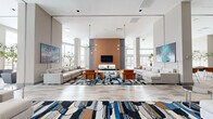 Located at 880 Pacific St in Stamford, Connecticut, this expansive single-floor amenity space offers a sophisticated blend of modern design and functional elegance. The layout encompasses several distinct areas unified by a consistent contemporary aesthetic, light-toned luxury vinyl and stone-look tile flooring, and soaring ceilings that contribute to an open, airy atmosphere throughout.

The central lounge is a showpiece, anchored by a warm terracotta accent wall housing a mounted flat-screen TV and fireplace. Symmetrically arranged neutral sectional sofas flank the room, complemented by cognac leather chairs and bold, abstract area rugs in blue, brown, and white. Dramatic sputnik pendant chandeliers hang overhead, while floor-to-ceiling windows flood the space with natural light and frame views of the surrounding Stamford skyline and outdoor pool terrace.

A curved glass curtain wall in a secondary lounge area offers panoramic outdoor vistas, creating a seamless connection between the interior and an expansive terrace. A transitional entry lounge with double-height ceilings and large glass entry doors provides a bright, welcoming arrival experience.

Additional highlights include a dedicated private screening room outfitted with leather reclining seats and acoustic wall panels, and a distinctive wine cellar featuring floor-to-ceiling lockable storage lockers, a rolling library ladder, and colorful bottle-shaped pendant lighting. A dining and social lounge with glass-wall separation and terrace access rounds out this well-appointed amenity floor.