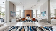 Located at 880 Pacific St in Stamford, Connecticut, this expansive single-floor amenity space offers a sophisticated blend of modern design and functional elegance. The layout encompasses several distinct areas unified by a consistent contemporary aesthetic, light-toned luxury vinyl and stone-look tile flooring, and soaring ceilings that contribute to an open, airy atmosphere throughout.

The central lounge is a showpiece, anchored by a warm terracotta accent wall housing a mounted flat-screen TV and fireplace. Symmetrically arranged neutral sectional sofas flank the room, complemented by cognac leather chairs and bold, abstract area rugs in blue, brown, and white. Dramatic sputnik pendant chandeliers hang overhead, while floor-to-ceiling windows flood the space with natural light and frame views of the surrounding Stamford skyline and outdoor pool terrace.

A curved glass curtain wall in a secondary lounge area offers panoramic outdoor vistas, creating a seamless connection between the interior and an expansive terrace. A transitional entry lounge with double-height ceilings and large glass entry doors provides a bright, welcoming arrival experience.

Additional highlights include a dedicated private screening room outfitted with leather reclining seats and acoustic wall panels, and a distinctive wine cellar featuring floor-to-ceiling lockable storage lockers, a rolling library ladder, and colorful bottle-shaped pendant lighting. A dining and social lounge with glass-wall separation and terrace access rounds out this well-appointed amenity floor.