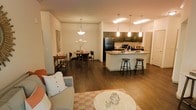 This single-floor residence showcases a thoughtfully designed open-concept floor plan that unifies the living, dining, and kitchen spaces into one cohesive and functional environment. Rich hardwood-style flooring flows throughout, lending warmth and continuity to the entire space.

The kitchen serves as a central hub, featuring dark cabinetry, a large island with bar seating for casual dining, pendant lighting, and black stainless steel appliances. The island provides generous counter space and encourages social interaction with adjacent living areas.

A dedicated dining area sits just off the kitchen, illuminated by an elegant pendant light fixture and comfortably accommodating a round dining table with seating for four. A side bar cart adds a stylish and practical touch to the space.

The living area is bright and inviting, furnished with a comfortable sofa and a round coffee table, anchored by a patterned area rug that adds color and texture. The open sightlines between all three zones create an airy, spacious atmosphere ideal for both everyday living and entertaining. Crown molding and neutral wall tones contribute to the clean, polished aesthetic throughout the space.