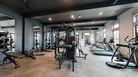 The fitness center at Evolve St. James Apartment Homes, located at 3025 Headwater Dr in Southport, NC, is a thoughtfully designed, single-floor facility offering a comprehensive workout experience across two dedicated exercise rooms connected by a hallway. The main exercise room features a dark, sophisticated aesthetic with deep charcoal-toned coffered ceilings, recessed lighting, and polished concrete-style flooring. This space is outfitted with a robust selection of strength training equipment, including multi-station cable machines, free weight racks with a full dumbbell set, and weight benches, all complemented by floor-to-ceiling mirrors that enhance the sense of space. A row of cardio machines — including treadmills, ellipticals, and a rowing machine — lines the windowed wall, allowing natural light to fill the room and offering outdoor views. The second exercise room provides a more boutique-style environment, distinguished by decorative patterned accent walls, elegant arched mirror details, and high ceilings. This room is dedicated to cycling and low-impact training, housing stationary bikes and spin cycles alongside a wall-mounted display screen and built-in cabinetry. Large windows throughout both rooms invite ample natural light, while the hallway entry features the branded Evolve St. James welcome mat and direct exterior access, providing a polished and functional transition between spaces.