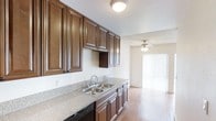 This single-floor space presents a well-appointed kitchen that seamlessly connects to an adjacent living area, creating an open and functional layout. The kitchen is outfitted with rich, dark-toned raised-panel cabinetry that extends to the upper walls, offering generous storage capacity. The countertops feature a granite-style surface in neutral tones, complementing the cabinetry and providing ample workspace. A double stainless steel sink is positioned beneath the upper cabinets, and a built-in dishwasher adds convenience. The flooring throughout transitions into warm wood-tone laminate, lending continuity between the kitchen and the adjoining living space. The living area benefits from abundant natural light streaming through a large sliding glass door dressed with vertical blinds, which also provides outdoor access. A ceiling fan with an integrated light fixture is centrally mounted in the living area, enhancing air circulation and overhead lighting. The overall ambiance is clean and neutral, with textured ceilings and light-toned walls that create a bright, welcoming atmosphere throughout the space.