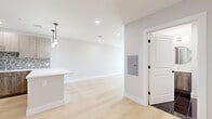 This contemporary single-floor residence showcases a thoughtfully designed open-concept layout with a clean, modern aesthetic throughout. Light-toned hardwood flooring runs seamlessly across the living areas, creating a bright and cohesive feel.

The kitchen is a standout feature, equipped with warm wood-grain cabinetry, white quartz countertops, a full suite of stainless steel appliances including a refrigerator, range, microwave, and dishwasher, and a distinctive hexagonal mosaic tile backsplash. A central island with pendant lighting adds both functionality and style, opening directly to the living area.

The living and bedroom spaces are bathed in natural light, courtesy of large black-framed windows that frame expansive views of the surrounding urban skyline. The crisp white walls and recessed lighting throughout lend a fresh, airy ambiance to every room.

The bathroom is finished with polished dark tile flooring and floor-to-ceiling white subway tile surrounding a full soaking tub, complemented by a round mirror and wood-toned vanity. A dedicated utility panel and well-placed storage solutions reflect careful attention to practical living. Overall, the space combines modern design with functional comfort in a bright, welcoming environment.