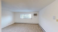This single-floor unit offers a practical, well-organized layout across several rooms, each finished with neutral tones and designed for comfortable everyday living. The spacious main living area features wall-to-wall beige carpeting, white walls, and a wide window fitted with blinds that allows natural light to filter in while providing views of surrounding greenery. A wall-mounted air conditioning unit and continuous baseboard heating ensure year-round climate control throughout the space.

The galley-style kitchen is efficiently arranged with light wood upper cabinetry, a white electric coil range, a full-size white refrigerator, and tile flooring. A ceiling fan overhead adds ventilation, and the layout opens toward a carpeted hallway connecting the rest of the unit.

The bedrooms are carpeted and feature ceiling fans with integrated lighting, windows with outdoor views, and baseboard heating. One bedroom includes an adjacent closet area, offering practical storage. The bathroom is neatly appointed with a combined tub and shower enclosed by a curtain, a standard toilet, and a charming mosaic tile floor in warm neutral tones. The overall ambiance of the unit is clean, functional, and move-in ready.