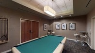 This single-floor recreational space is thoughtfully designed to offer a refined and inviting atmosphere for leisure activities. At the heart of the room sits a full-sized billiards table with a vibrant teal felt surface, serving as the focal point of the space. The room is finished with warm taupe walls that create a sophisticated, contemporary ambiance throughout.

Overhead, a large rectangular pendant light fixture with a neutral fabric shade provides soft, even illumination, complemented by recessed ceiling lights that enhance the room's brightness. The ceiling features a subtle raised tray detail, adding an architectural element that lends a sense of depth and elegance.

Along one wall, a set of rich wood-paneled double doors offer a stylish entryway, while a small bistro-style table with modern curved chairs provides a casual seating area for guests not at the table. A series of framed black-and-white portrait photographs in matching silver frames lines another wall, adding a curated, gallery-like quality to the decor.

The flooring transitions between materials, adding visual interest and defining areas within the space. Overall, this room balances functionality with polished design, making it an ideal setting for social entertainment and relaxed recreation.