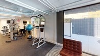 Located in Los Angeles, CA 90024, this fitness center offers a comprehensive workout facility spread across a single level. The space is designed with functionality in mind, featuring an exposed ceiling with visible ductwork and piping, accented by linear fluorescent lighting that ensures a bright, well-lit environment throughout.

The cardio zone is equipped with multiple treadmills, a spin bike, and a stationary upright bike, each positioned alongside wall-mounted televisions for an engaging workout experience. Rubber flooring spans the exercise areas, providing durability and comfort underfoot.

The strength training section houses a multi-station cable machine, a power tower for pull-ups and dips, and a dedicated free weights area. Full-length mirrors along one wall visually expand the space and allow for proper form checks during workouts. Stability balls and additional accessories are also available for functional training.

A transitional lounge area near the entrance features stylish leather Barcelona-style chairs set on a patterned tile floor, offering a relaxed space to rest. Large sliding glass doors open directly to an outdoor pool area, flooding this zone with natural light and providing a seamless indoor-outdoor connection. The overall ambiance is clean and utilitarian, with a modern aesthetic suited for a focused fitness environment.