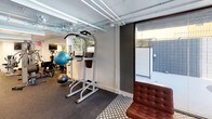 Located in Los Angeles, CA 90024, this fitness center offers a comprehensive workout facility spread across a single level. The space is designed with functionality in mind, featuring an exposed ceiling with visible ductwork and piping, accented by linear fluorescent lighting that ensures a bright, well-lit environment throughout.

The cardio zone is equipped with multiple treadmills, a spin bike, and a stationary upright bike, each positioned alongside wall-mounted televisions for an engaging workout experience. Rubber flooring spans the exercise areas, providing durability and comfort underfoot.

The strength training section houses a multi-station cable machine, a power tower for pull-ups and dips, and a dedicated free weights area. Full-length mirrors along one wall visually expand the space and allow for proper form checks during workouts. Stability balls and additional accessories are also available for functional training.

A transitional lounge area near the entrance features stylish leather Barcelona-style chairs set on a patterned tile floor, offering a relaxed space to rest. Large sliding glass doors open directly to an outdoor pool area, flooding this zone with natural light and providing a seamless indoor-outdoor connection. The overall ambiance is clean and utilitarian, with a modern aesthetic suited for a focused fitness environment.