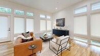 Located at 5021 Bryan St in Dallas, TX 75206, this modern single-story unit offers a thoughtfully designed open-concept layout finished with warm hardwood flooring throughout. The combined kitchen and living area forms the heart of the space, where soaring ceilings and a generous bank of floor-to-ceiling windows with transom panes flood the room with natural light, creating an airy and inviting atmosphere. The living area is spacious enough to accommodate comfortable seating arrangements and benefits from direct access to the outdoors. The kitchen is outfitted with sleek white flat-front cabinetry, white quartz countertops, a large center island with bar seating, and a full suite of stainless steel appliances including a French door refrigerator, range, and built-in microwave. Recessed lighting enhances the clean, contemporary aesthetic. The bathroom is a standout feature, showcasing a walk-in glass-enclosed shower clad in sage green subway tile, a white floating vanity with a quartz countertop, and penny-tile flooring that adds a refined textural detail. The dedicated closet provides practical storage with built-in hanging rods and continues the hardwood flooring found throughout the unit. The overall design palette of crisp whites, natural wood tones, and green accents creates a cohesive, modern aesthetic ideal for East Dallas living.
