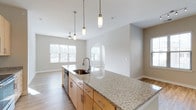 This single-floor, 2-bedroom with den residence features an open-concept design that seamlessly connects the kitchen and living areas. The kitchen is a standout, anchored by a large center island topped with speckled granite countertops that provide generous prep and entertaining space. A stainless steel undermount sink with a modern gooseneck faucet is integrated into the island, while pendant lights with brass-toned fixtures hang overhead, adding a warm, contemporary accent. Light maple cabinetry lines the perimeter walls, complemented by stainless steel appliances including a refrigerator, range, microwave, and dishwasher. The flooring throughout is a light-toned wood-style material that contributes to the bright, airy atmosphere. The living area flows naturally from the kitchen and receives ample natural light through multiple windows with horizontal blinds. A ceiling fan with a light fixture is centrally positioned in the living space. A distinctive arched architectural niche adds character to the transition between spaces. The neutral color palette of soft whites and grays throughout creates a clean, cohesive aesthetic well-suited to a variety of furnishing styles.