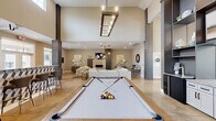 Located at 1000 Sweet Almond Dr in Holly Springs, NC, this generously sized single-floor community amenity space offers a well-rounded mix of social, recreational, and fitness facilities. The open-concept main area makes an immediate impression with soaring ceilings, dramatic dark accent columns, and polished concrete floors that extend throughout. A statement billiards table anchors the central gathering space, illuminated by a striking rectangular pendant light fixture overhead. Adjacent to it, a comfortable living room features a large sectional sofa, fireplace with a tile surround, mounted television, and stylish pendant lighting — ideal for relaxed socializing. The kitchen and dining area are equipped with granite countertops, gray cabinetry, a coffee station, and bistro-style high-top seating. A separate dining nook with a chandelier and large windows invites natural light, while decorative wall art adds warmth throughout. The dedicated exercise room is well-appointed with strength training equipment, cardio machines, a punching bag, mirrored walls, and ceiling fans — all bathed in natural light from oversized windows. A connected spin/yoga studio offers additional workout versatility with hardwood-style flooring. Rounding out the space, a covered outdoor patio provides comfortable lounge seating with direct views of the community pool.