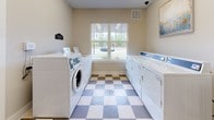 Located at 3500 Pinckney Marsh Ln in Summerville, South Carolina, this single-floor community laundry facility is designed with both functionality and comfort in mind. The space is finished with a distinctive two-tone checkered tile floor in neutral tan and cream tones, lending a clean and classic aesthetic throughout.

The laundry area is well-appointed with multiple front-loading and top-loading washers and dryers arranged along the walls, offering ample capacity for residents. A large window on one wall floods the space with natural light and provides a view of the surrounding greenery and parking area, creating a bright and pleasant environment. Overhead lighting further ensures the room is well-illuminated throughout the day and evening.

The room includes practical amenities such as a wall-mounted flat-screen television, a seating bench for waiting residents, a small utility table, and a stainless steel waste bin. An electrical panel access box is conveniently recessed into the wall. Entry is provided through a dark-framed glass-paned door that connects to an adjacent space, as well as a secondary white interior door, allowing for easy access and circulation.

The neutral, light-colored walls and clean finishes create an organized and inviting atmosphere, making this community laundry room a practical and comfortable shared amenity.