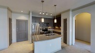 This single-floor residence offers a thoughtfully designed open-concept layout that seamlessly blends functionality with modern aesthetics. The kitchen serves as the heart of the space, showcasing rich dark cabinetry that extends to the ceiling for generous storage capacity. A central island with a granite countertop and an integrated double sink acts as both a practical workspace and a natural gathering point. Stainless steel appliances, including a refrigerator, range, and built-in microwave, provide a sleek, contemporary finish. The kitchen is illuminated by a combination of elegant cylindrical pendant lights hanging above the island and a track lighting system overhead, creating a warm and inviting atmosphere. A tasteful tile backsplash adds texture and depth to the cooking area. Neutral-toned carpeting flows throughout the surrounding living areas, complementing the overall color palette of soft grays and warm whites. Architectural column details and clean door moldings contribute a refined, structured character to the space. The open layout enhances the sense of spaciousness, making the home feel bright, welcoming, and well-appointed throughout.