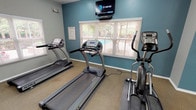 Located at 6300 N MacArthur Blvd in Irving, Texas, this single-floor fitness center is designed to provide residents with a functional and well-equipped workout facility. The space is organized into distinct exercise zones, each thoughtfully appointed to support a range of fitness activities.

The cardio area features multiple treadmills and an elliptical trainer, arranged along a teal-accented wall beneath a wall-mounted television. Natural light filters in through several windows dressed with horizontal blinds, offering views of the outdoor pool and surrounding greenery. A ceiling fan overhead provides additional air circulation during workouts.

Adjacent to the cardio zone, a dedicated strength training area houses a multi-station cable and weight machine, complemented by a flat bench. A large floor-to-ceiling mirror spans one wall of this section, allowing users to monitor their form while exercising and visually expanding the sense of space.

The entryway and transitional corridor feature durable luxury vinyl plank flooring, neutral gray walls, and fluorescent lighting, providing a clean and practical pathway through the facility. Additional doors offer convenient access to the exterior of the building. The textured ceiling and consistent, well-maintained finishes throughout contribute to the overall tidy and functional ambiance of this community fitness center.