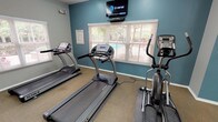Located at 6300 N MacArthur Blvd in Irving, Texas, this single-floor fitness center is designed to provide residents with a functional and well-equipped workout facility. The space is organized into distinct exercise zones, each thoughtfully appointed to support a range of fitness activities.

The cardio area features multiple treadmills and an elliptical trainer, arranged along a teal-accented wall beneath a wall-mounted television. Natural light filters in through several windows dressed with horizontal blinds, offering views of the outdoor pool and surrounding greenery. A ceiling fan overhead provides additional air circulation during workouts.

Adjacent to the cardio zone, a dedicated strength training area houses a multi-station cable and weight machine, complemented by a flat bench. A large floor-to-ceiling mirror spans one wall of this section, allowing users to monitor their form while exercising and visually expanding the sense of space.

The entryway and transitional corridor feature durable luxury vinyl plank flooring, neutral gray walls, and fluorescent lighting, providing a clean and practical pathway through the facility. Additional doors offer convenient access to the exterior of the building. The textured ceiling and consistent, well-maintained finishes throughout contribute to the overall tidy and functional ambiance of this community fitness center.