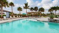 Located at 28790 Versol Dr in Bonita Springs, Florida, this outdoor amenity complex delivers a resort-style experience designed for relaxation and recreation. The centerpiece of the space is a series of sparkling, large-format swimming pools with crystal-clear turquoise water, each surrounded by expansive paver pool decks ideal for sunbathing and socializing. Depth markers are clearly posted throughout, and stainless steel entry rails provide convenient pool access.

Tall royal palms and sabal palms are strategically planted throughout the grounds, creating a lush tropical canopy that provides both shade and visual appeal. A beautifully crafted pergola with an open-beam roof serves as a covered lounge area, furnished with comfortable seating and accented by colorful tropical plantings. Teal-accented umbrellas and lounge chairs are arranged around the pool decks, offering shaded retreats for guests.

A stone waterfall feature flows into one of the pools, adding a distinctive natural design element. An outdoor kitchen or grilling station is also visible, enhancing the entertaining capabilities of the complex. The adjacent clubhouse building features a metal roof and covered porch with additional seating. The overall ambiance is bright, airy, and quintessentially Floridian, making this a standout communal outdoor destination.