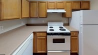 This single-floor space is designed with a practical and straightforward layout that prioritizes functionality. The kitchen is equipped with warm honey-toned wood cabinetry offering generous upper and lower storage, light-colored countertops, a coil-burner electric range with overhead ventilation, and white appliances including a dishwasher and refrigerator. A peninsula counter connects the kitchen to the main living area, creating an open-concept flow that enhances the sense of space.

The living area is carpeted in neutral beige tones and provides an open, versatile space suited for various furniture arrangements. A front entry door with hardwood flooring inset leads directly into this area, adding a functional transition zone. The bedroom is similarly carpeted, featuring neutral walls, a six-panel white door, and a window that allows natural light to filter in.

The bathroom features warm hardwood flooring, a full tub-shower combination with white tile surround and grab bars for accessibility, a toilet, a vanity with under-sink storage, and an overhead wall cabinet. Throughout the unit, neutral wall tones, white trim, and consistent finishes contribute to a clean, cohesive aesthetic.