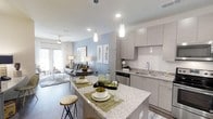 This single-floor residence showcases a thoughtfully designed open-concept layout that seamlessly connects the kitchen and living areas, creating a bright and cohesive living environment. The contemporary kitchen is outfitted with sleek, flat-front cabinetry in a warm gray tone, gleaming granite countertops, and a large central island that doubles as a breakfast bar with seating. A white subway tile backsplash adds a clean, modern touch, while stainless steel appliances complete the culinary space. Elegant pendant lights hang above the island, providing focused task lighting and a refined aesthetic accent. The living area flows naturally from the kitchen, featuring a soothing blue accent wall that anchors the space and adds a layer of visual depth. Large windows and glass doors allow ample natural light to fill the room, while a ceiling fan ensures year-round comfort. Durable wood-style flooring runs throughout, tying the spaces together with warmth and continuity. The overall ambiance is modern and inviting, with a neutral color palette punctuated by carefully chosen décor, making this space both functional and visually appealing for everyday living and entertaining alike.